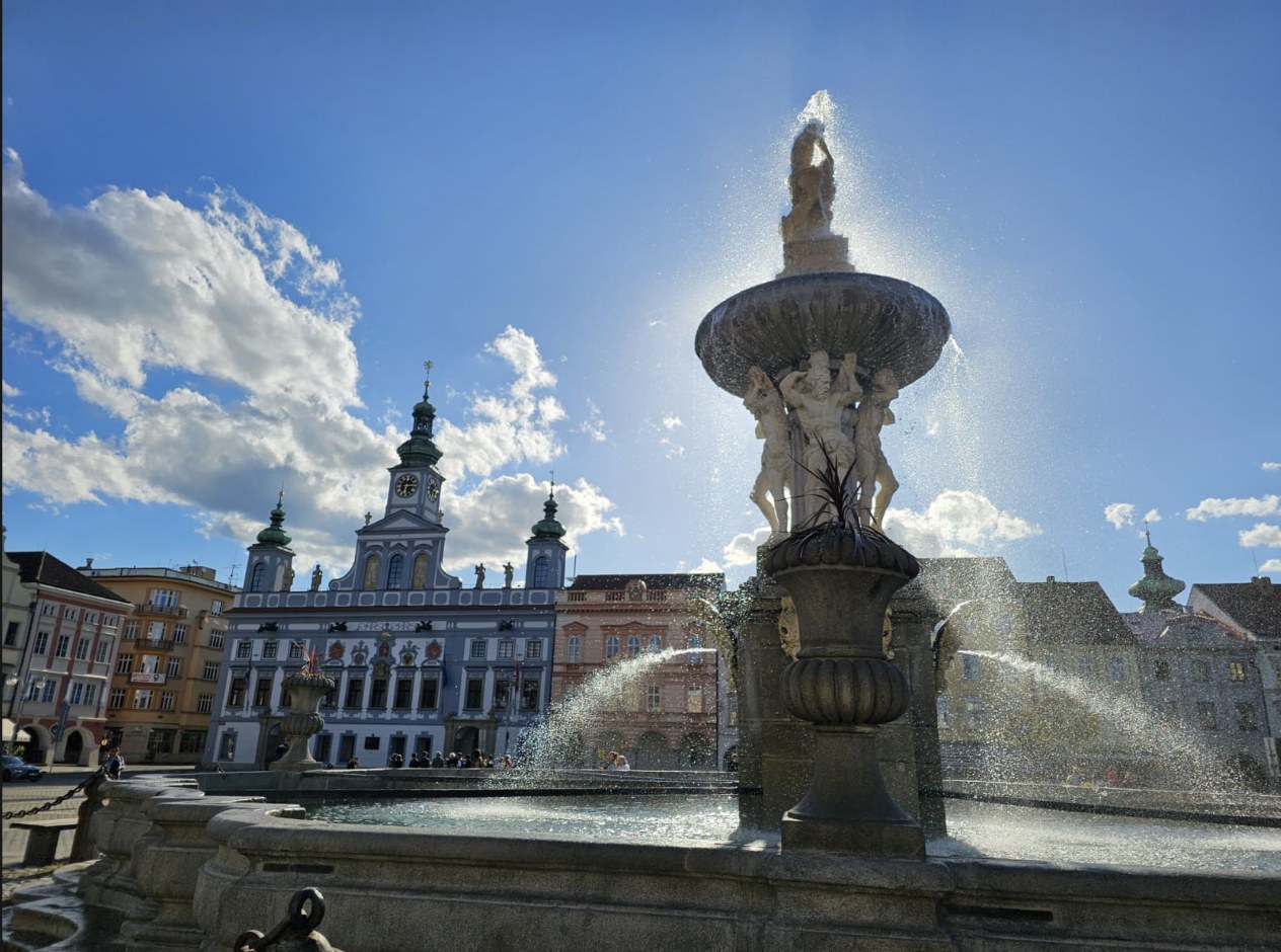 Fuente barroca en primer plano y el ayuntamiento de České Budějovice al fondo, bajo un cielo azul
