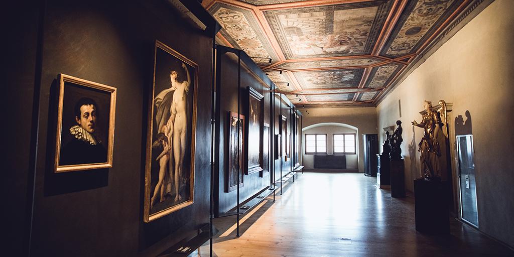 Sala de galería con retratos y pinturas antiguas en las paredes, esculturas doradas y techo artesonado pintado