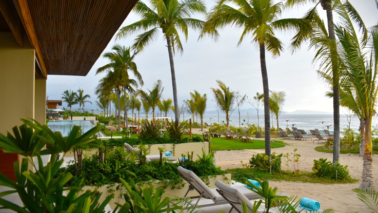 Jardín de un resort con palmeras, alberca y camastros frente a una playa de arena, con el mar y las montañas al fondo