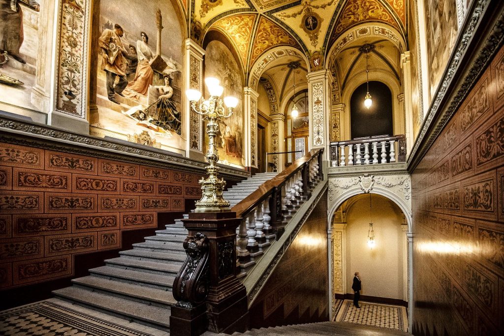 Escalinata monumental con barandilla de balaustres, lámpara de bronce, murales y bóvedas decoradas en el interior de un museo
