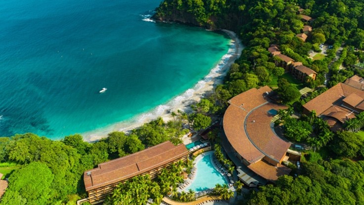 Vista aérea de un resort de edificios con tejados de teja entre vegetación tropical, junto a una bahía de aguas turquesas y una playa