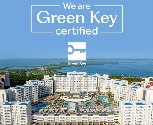 Vista aérea de un resort frente al mar, con edificios blancos y azules, junto a la leyenda «We are Green Key certified» y el logotipo de Green Key