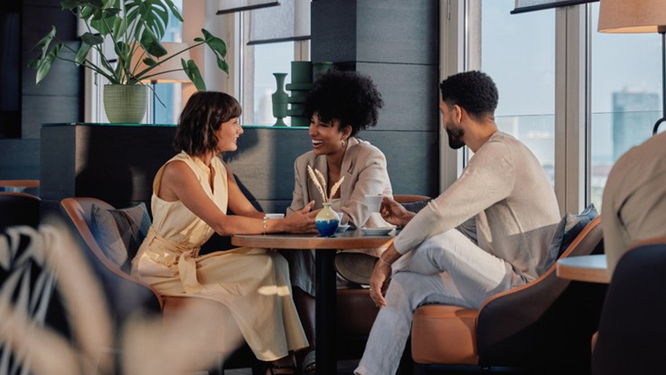 Tres personas conversan sentadas alrededor de una mesa en un salón de hotel con grandes ventanales