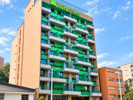 Fachada de un edificio de ladrillo con balcones verdes y el letrero ibis Styles en la parte superior, bajo un cielo azul.