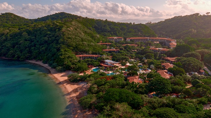Vista aérea del resort con edificios de tejas rojas entre vegetación tropical, junto a una bahía de aguas turquesas y una playa