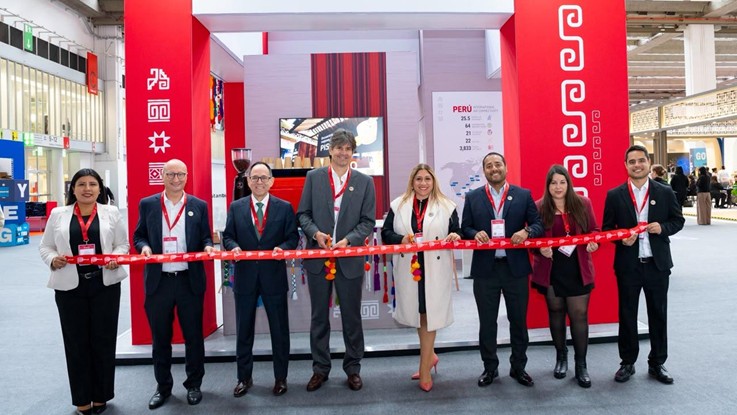 Grupo de personas con credenciales cortando una cinta roja frente a un stand con motivos rojos y un panel con el nombre Perú
