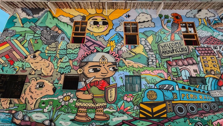 Mural colorido en la boletería de PeruRail en Machupicchu con un tren azul, un oso de anteojos, un personaje inca, el sol y el letrero Welcome to Machupicchu
