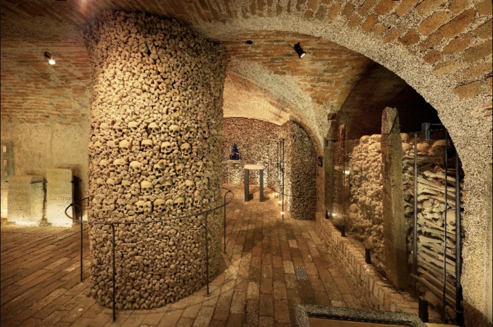 Galería subterránea con columnas y muros formados por miles de cráneos y huesos humanos iluminados con luz cálida