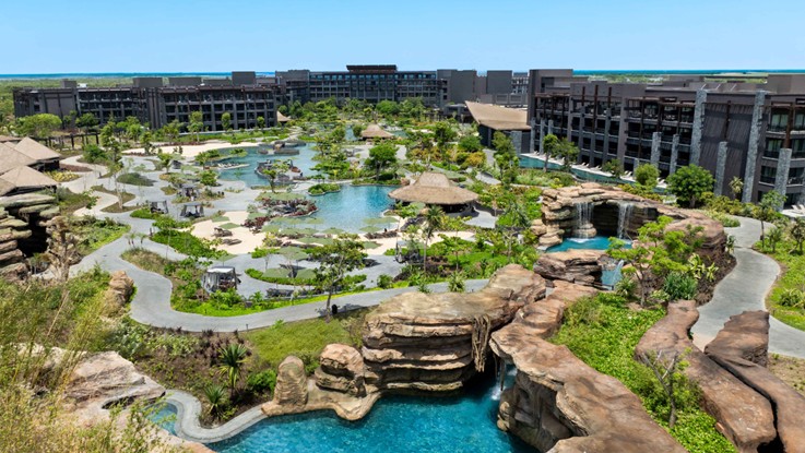 Vista aérea de un resort con una gran laguna, piscinas con cascadas de roca, jardines y edificios al fondo.