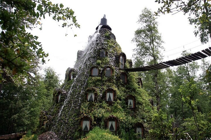Edificio en forma de montaña cubierto de vegetación, con una cascada y una pasarela colgante entre el bosque