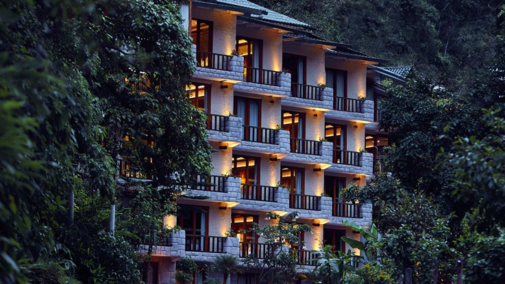 Fachada de un hotel de varias plantas con balcones iluminados, rodeada de vegetación espesa, al atardecer