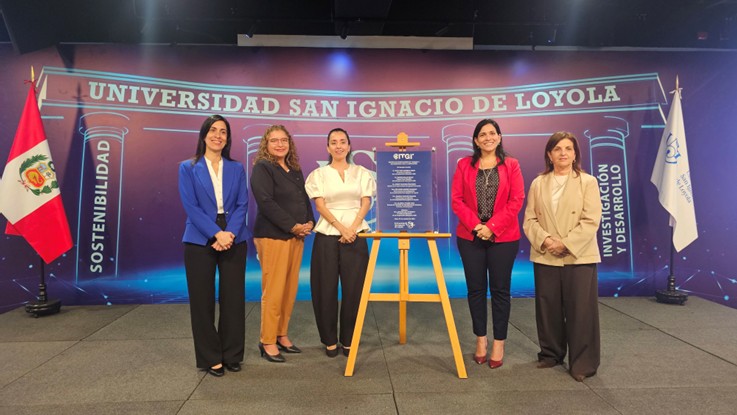 Cinco mujeres posan junto a una placa sobre un caballete, frente a un fondo con el nombre de la Universidad San Ignacio de Loyola y las banderas de Perú y de la universidad